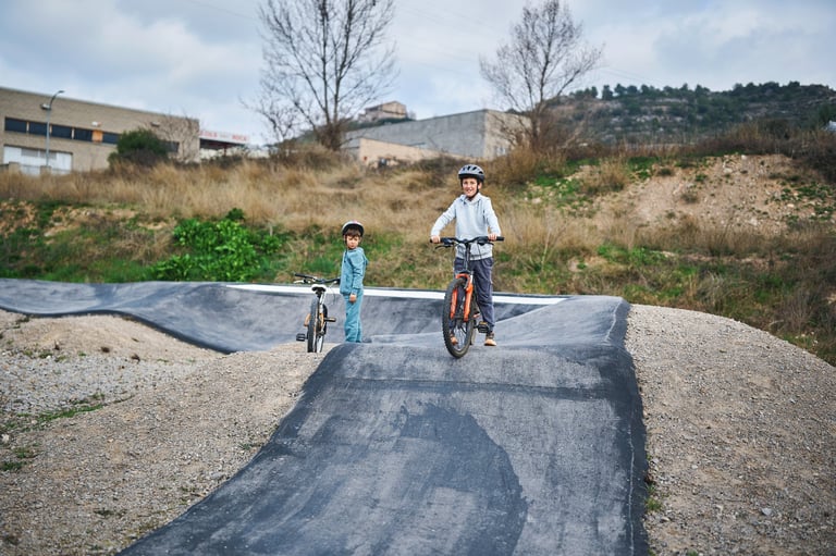 Two brothers with bicycles on an outdoor pump track, the older child ready to ride while the younger sibling waits behind