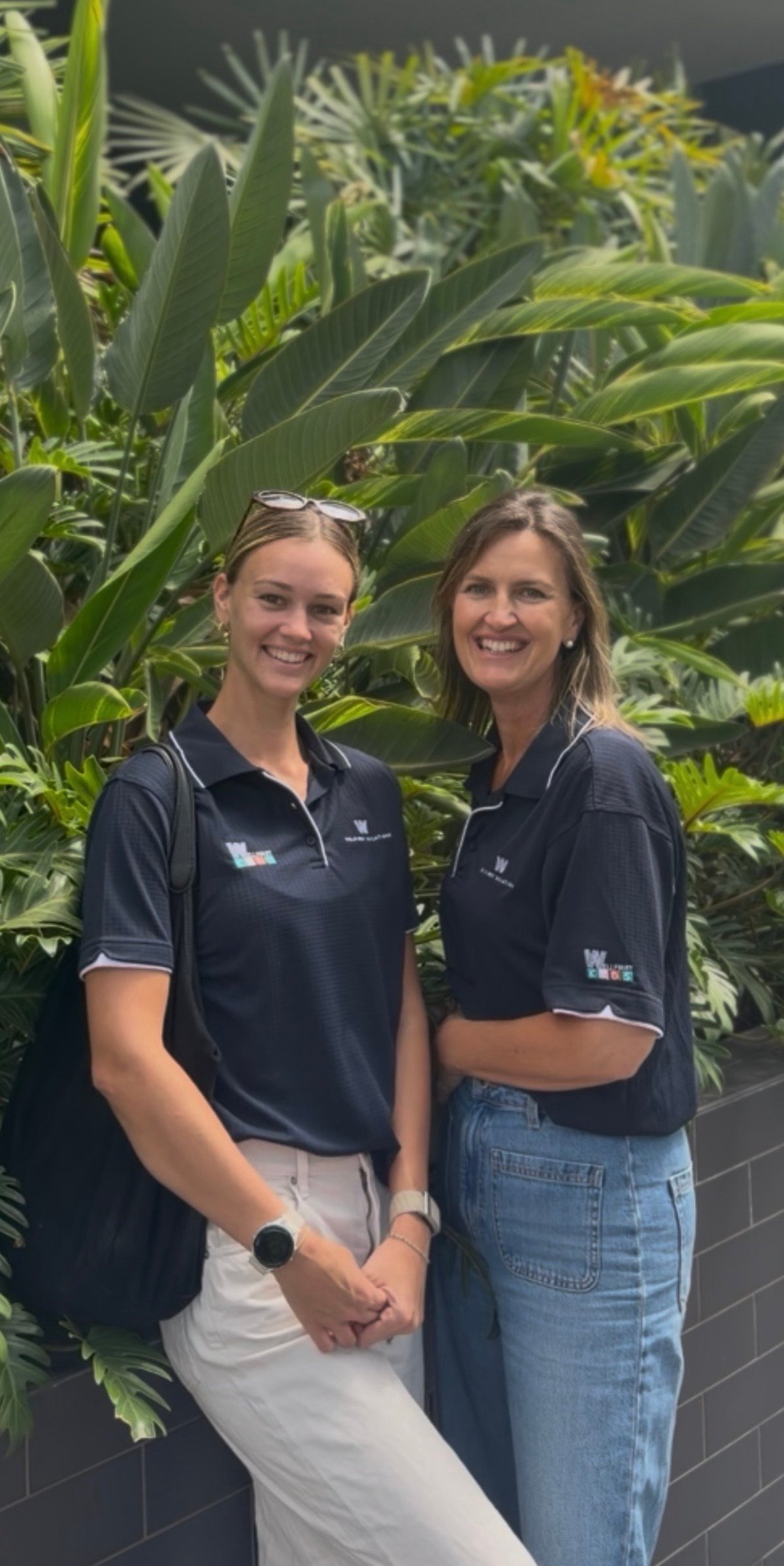 Two women in navy polo shirts posing together in front of tropical green plants and foliage